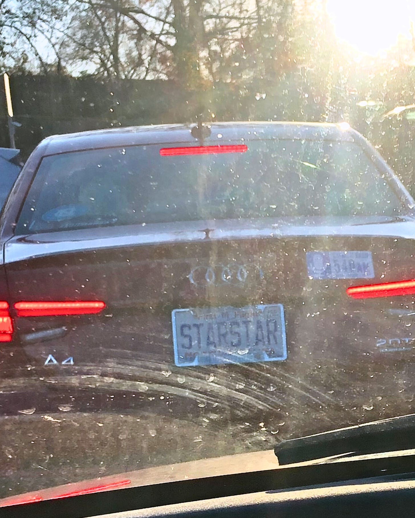 image taken from behind the wheel looking out of a dirty windshield at sunset toward the car in front which has a license place that can just barely be seen that says “STARSTAR”