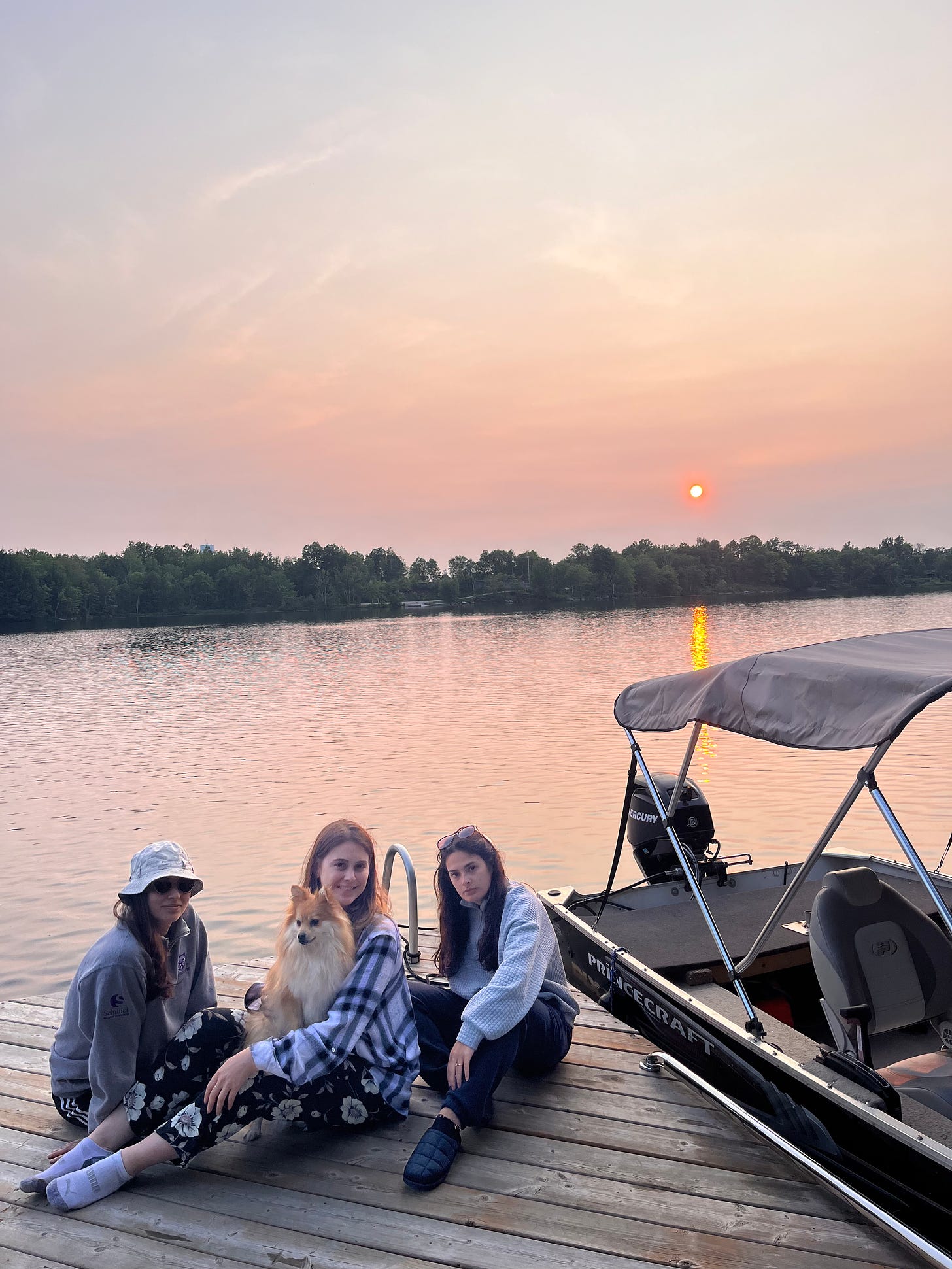 Three girls sitting on a wooden dock beside a boat. Sunset over lake in the background.