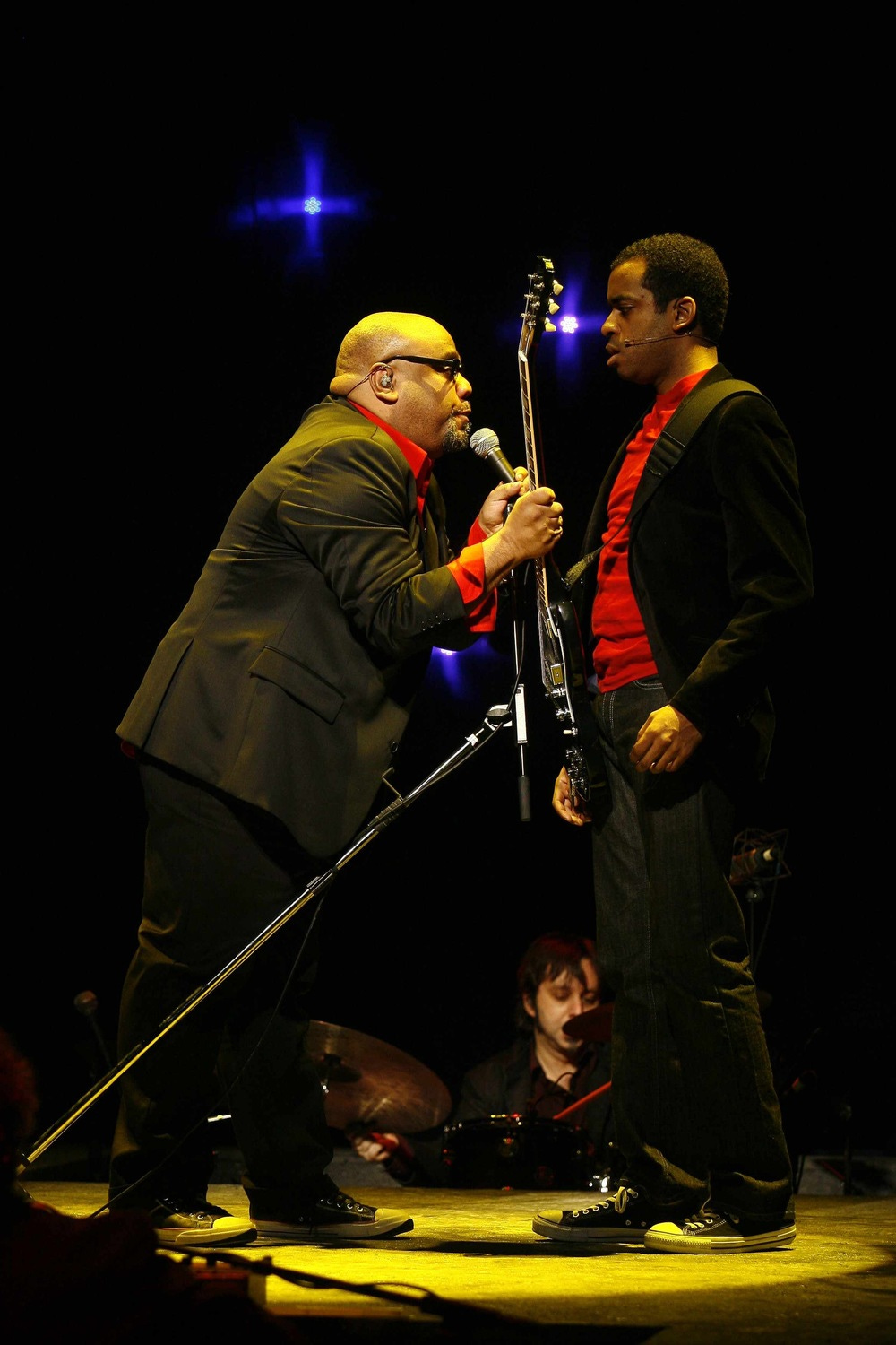 A man in a suit and glasses leans over a microphone towards a young man with a guitar