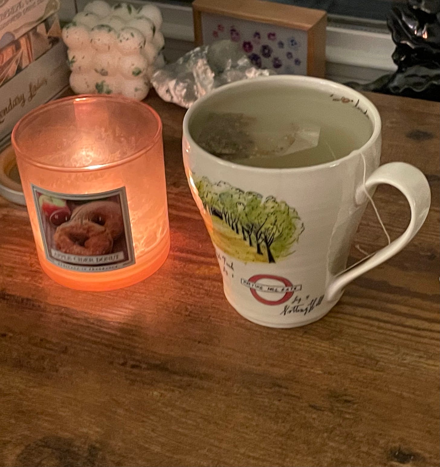 A candle named Apple Cider donut burns while sitting next to a London themed mug which is steeping tea.