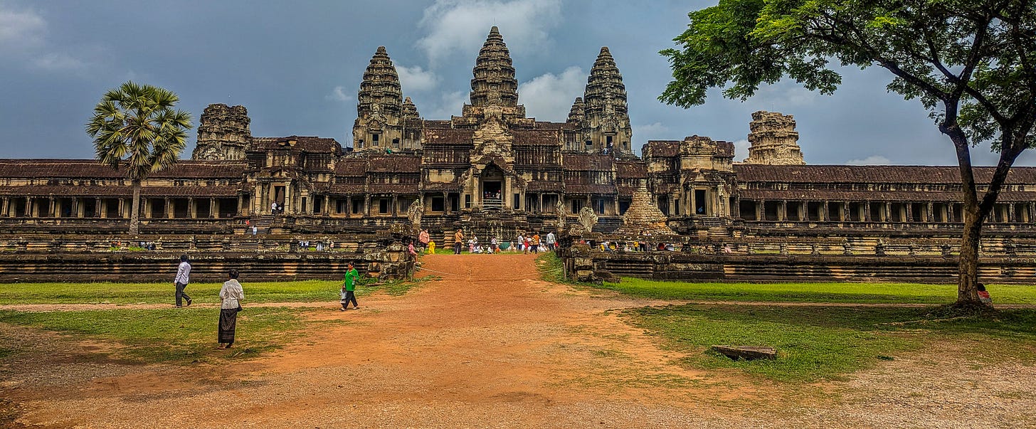 View of Angkor Wat approaching on a dirt path, three of the lotus-shaped temples framed in the center. 