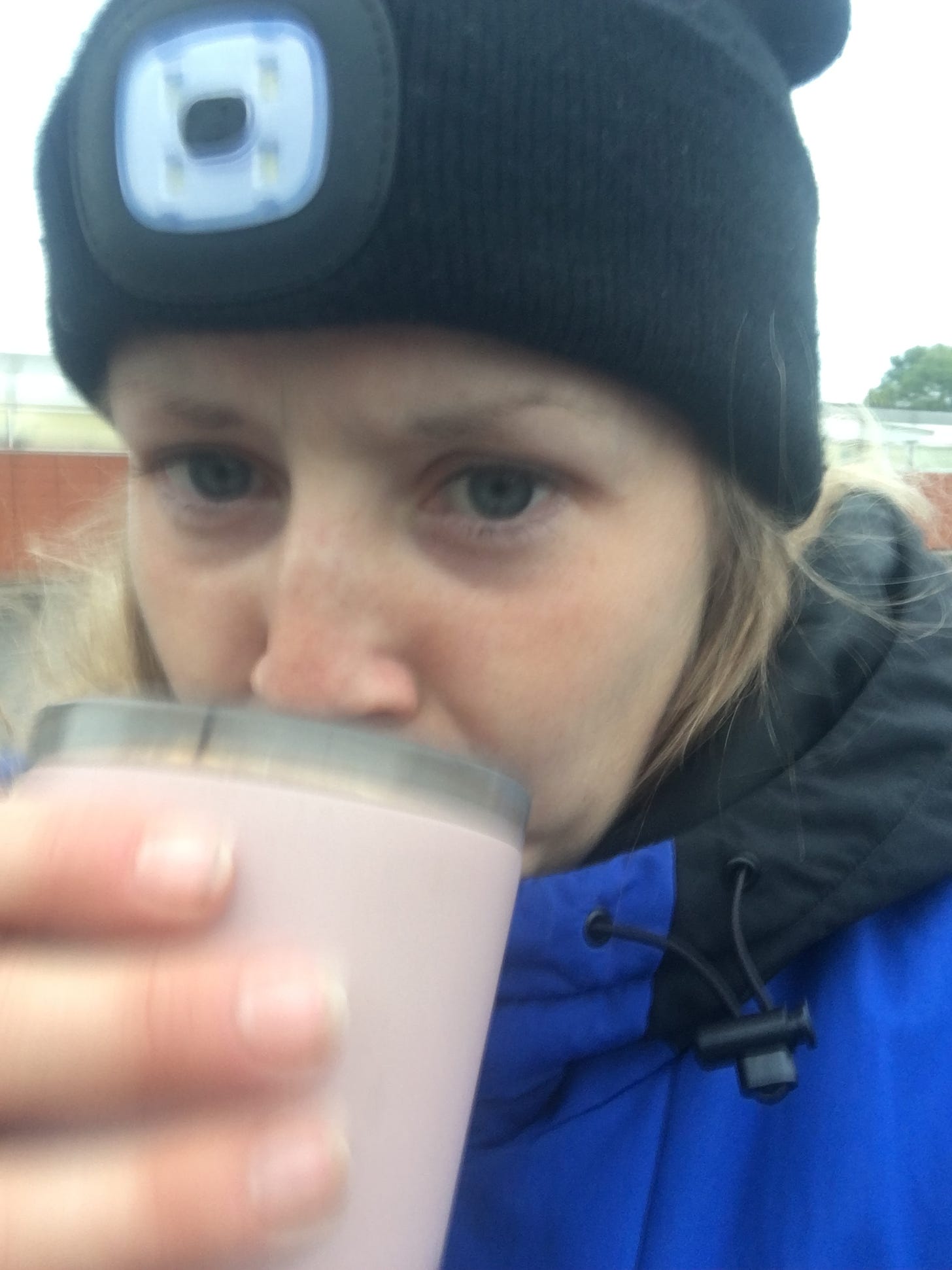 White woman wearing a hat with a light on the front and a puffer jacket sips a cup of tea.