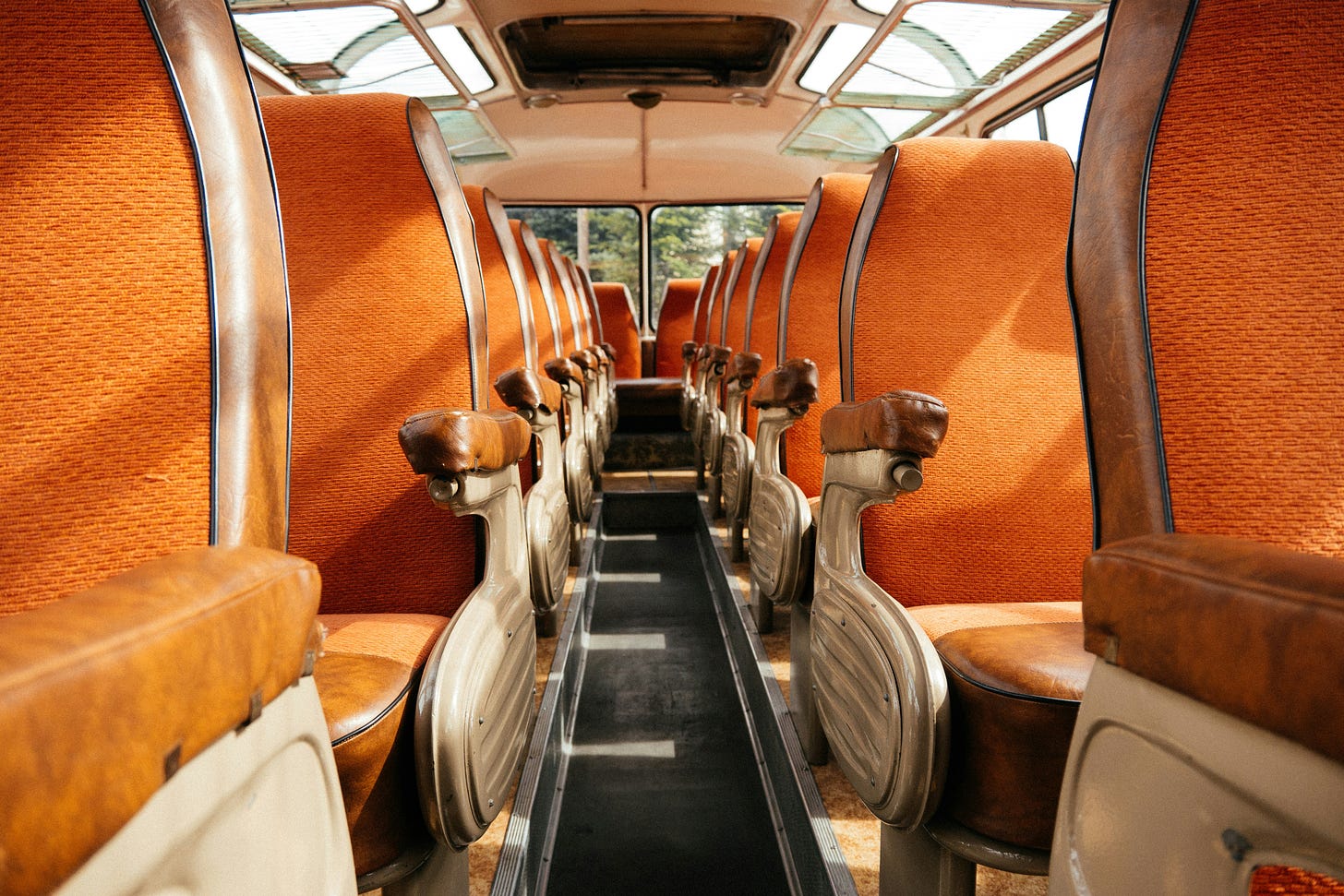 Rows of empty bus seats, in orange fabric. At the back, a view of trees is seen out the back window.