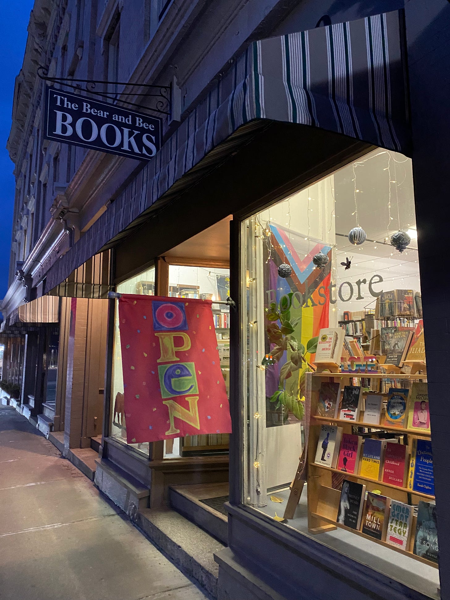 A lit up bookstore window on a dark night. There’s a colorful display of books and a big pride flag.