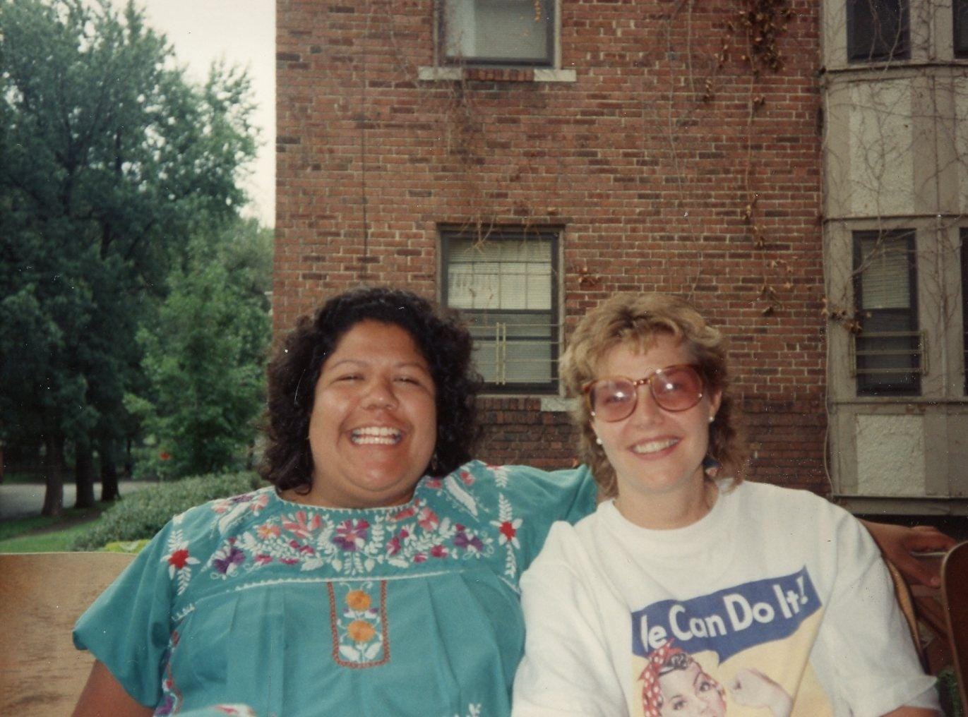 A Chicana woman wearing a traditional embroidered turquoise shirt, with a radiant smile on her face ringed by curly brown hair. She sits with her arm draped over the shoulders of a white blonde woman wearing a 'We Can Do It!' t-shirt and beige tinted glasses.