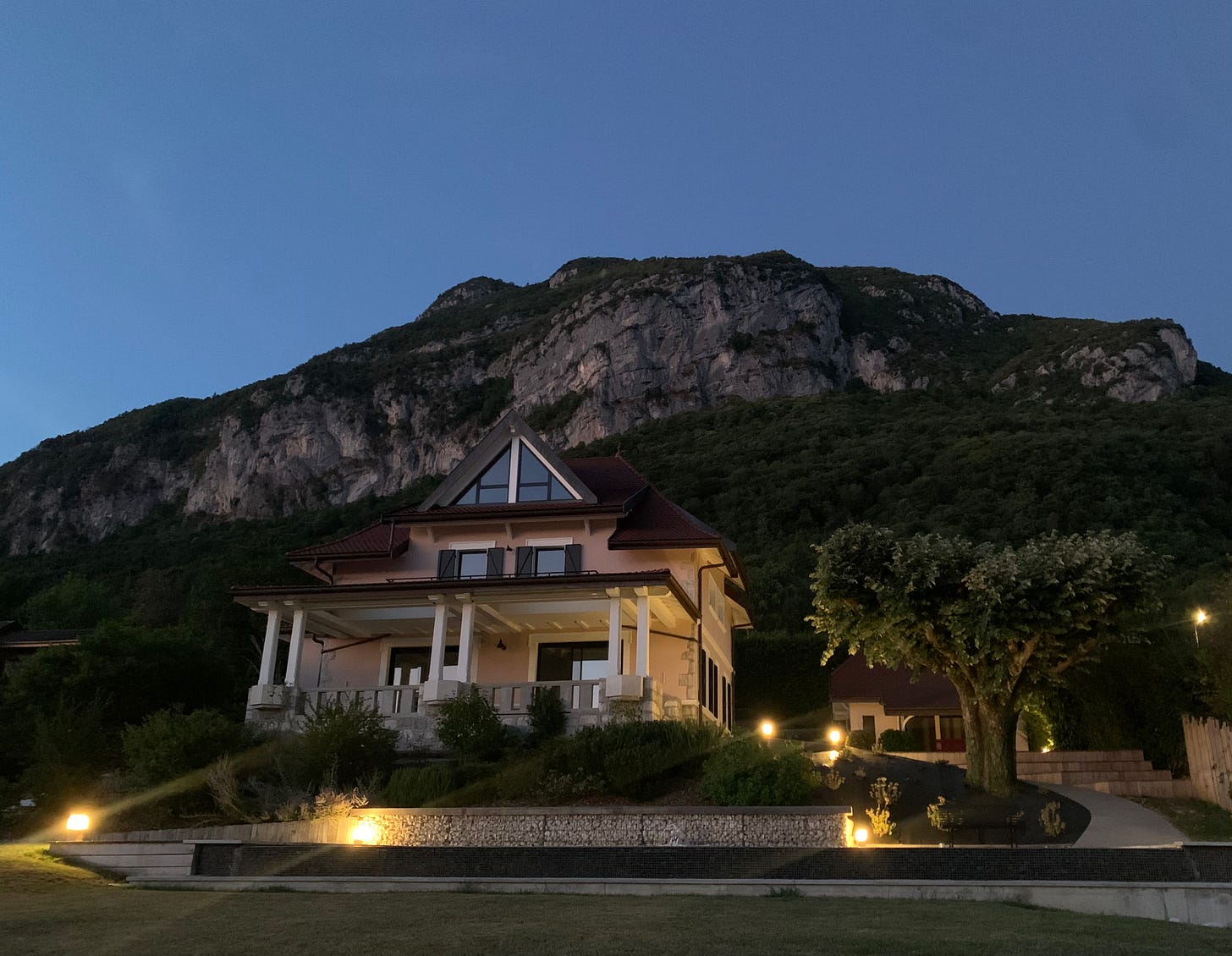 "A beautiful home in Veyrier-du-Lac, France, set against the backdrop of a rocky hillside and lush greenery, illuminated by outdoor lights during twilight."