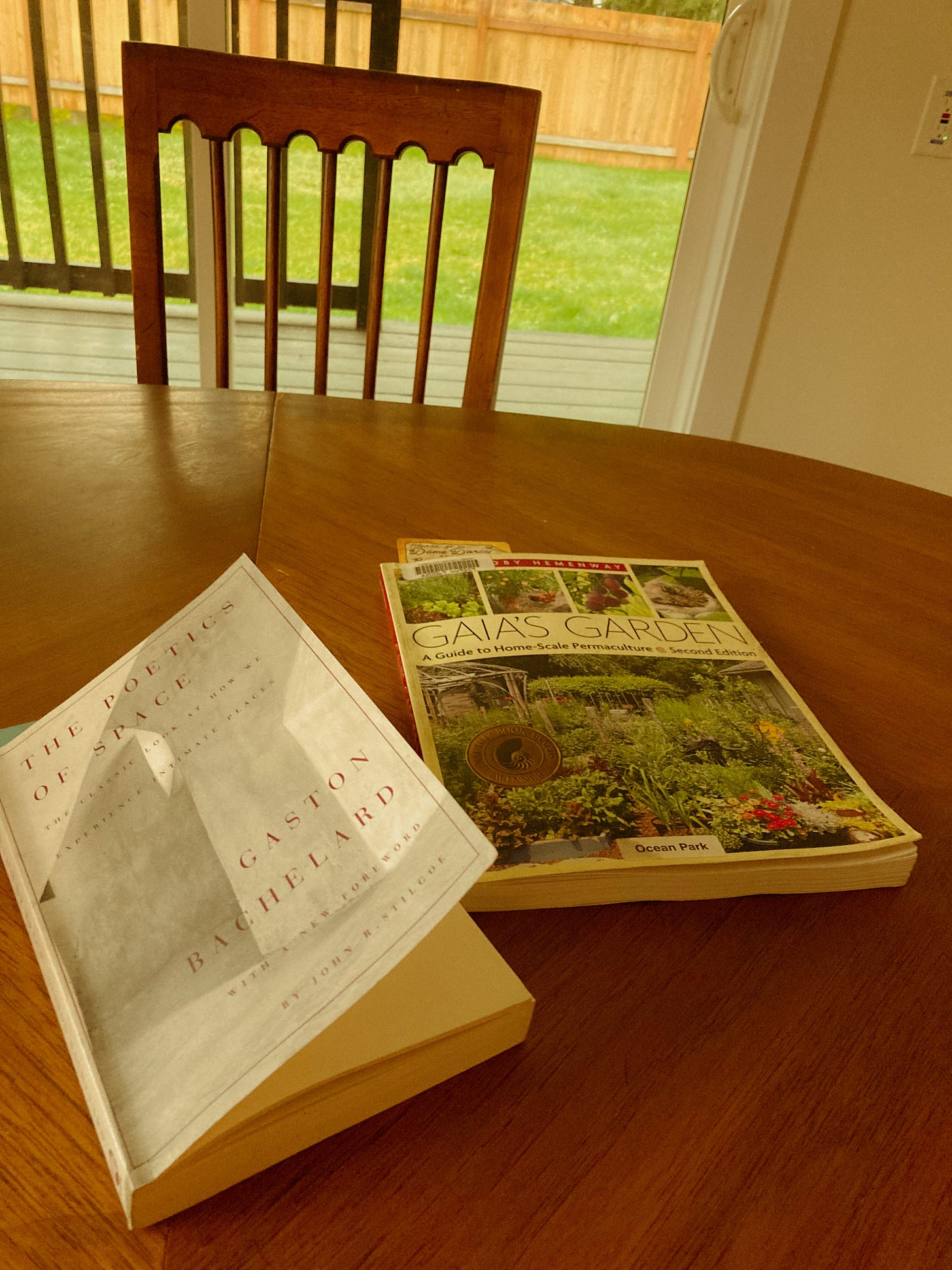 The books The Poetics of Space and Gaia's Garden lay on top of a kitchen table, in front of a sliding door which looks out at a grassy lawn.