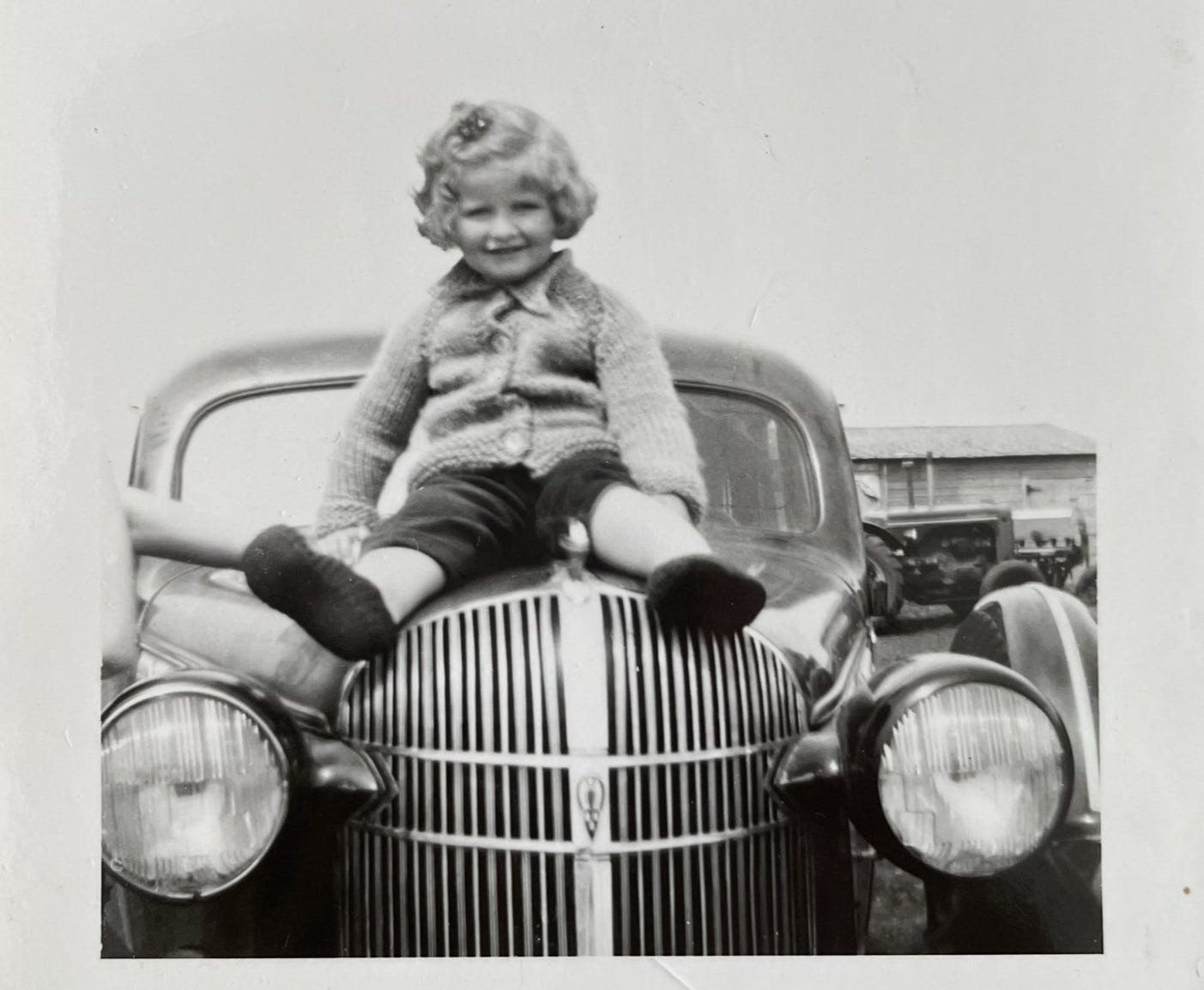 A black and white photo showing a young girl smiling from the hood of a 1940s sedan.