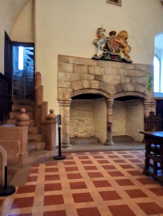 Carpeted room with coat of arms over double fireplace, entrance from visible spiral staircase on left