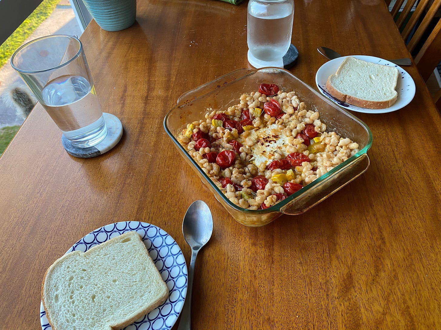 A square pyrex dish of beans, grape tomatoes, and yellow zucchini, with feta covered in chili flakes in the centre. Two small plates on either side have a slice of sourdough bread on them and a spoon beside.