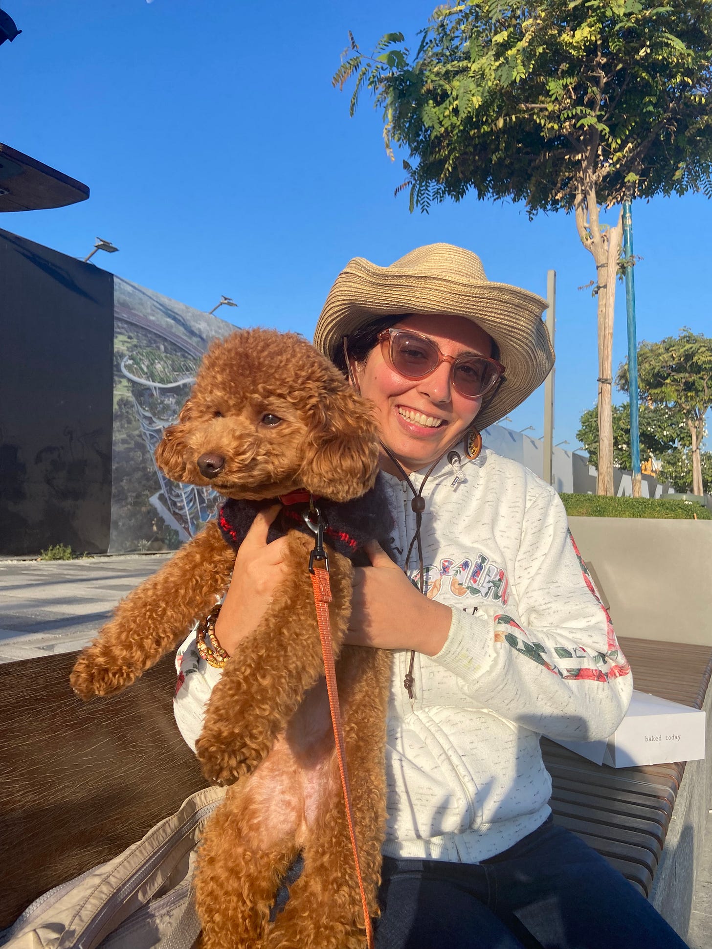Photo of a Middle Eastern woman sitting on a bench outside in a tree-lined walkway during a sunny morning while holding a small brown dog in her hands on its hindlegs. She is wearing a pair of jeans, a white sweatshirt with colorful print on the chest and sleeves, a pair of sunglasses, and a hat with a wide smile. Photo of a Middle Eastern woman sitting on a bench outside in a tree-lined walkway during a sunny morning while holding a small brown dog in her hands on its hindlegs. She is wearing a pair of jeans, a white sweatshirt with colorful print on the chest and sleeves, a pair of sunglasses, and a hat with a wide smile.
