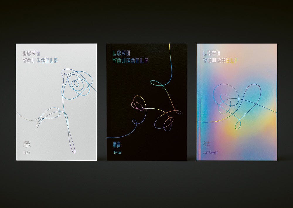 aerial view of 3 album covers titled "love yourself". Left to right they are white, black, and irridescent, each with a thin line illustrations
