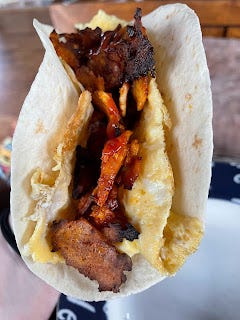 A tortilla wrap, held open so the filling can be seen. Visible in the photo is what looks like crispy pieces of bacon. They're nestling in a lining of omelette. There are a few splashes of tomato ketchup over everything.
