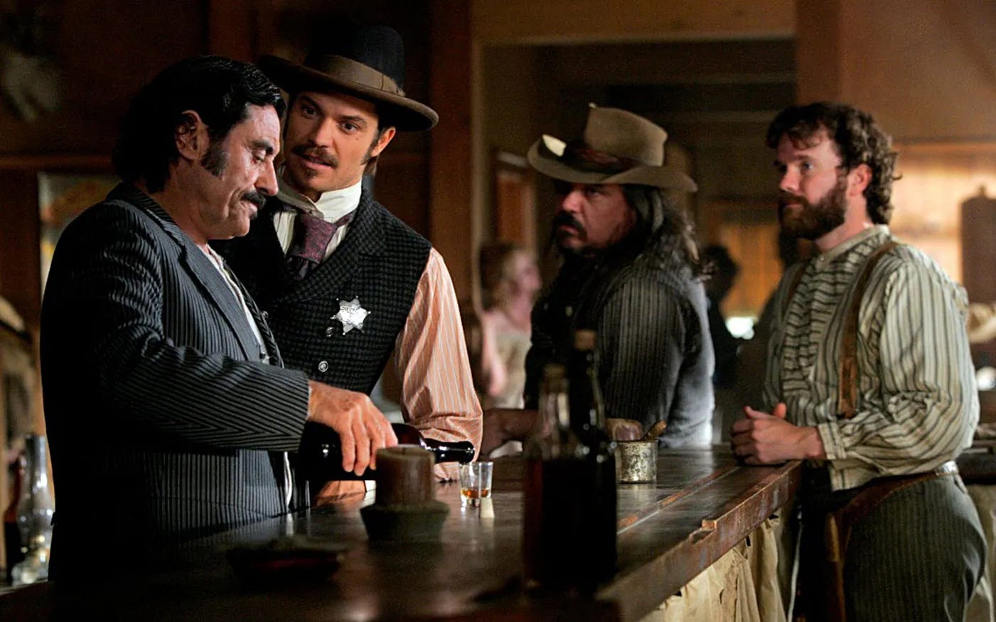 This image shows four men standing at the Gem Saloon's bar in a scene from David Milch's television series Deadwood. From left to right: Al Swearengen (played by Ian McShane, pouring whiskey into a shot glass), Sheriff Seth Bullock (played by Timothy Olyphant), Dan Dority (played by W. Earl Brown), and Johnny Burns (played by Seth Bridgers).