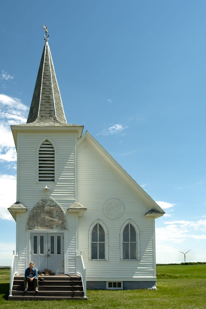 A white woman with pulled-back blond hair wearing a denim shirt sits on the front steps of an abandoned church with a tall steeple in Columbus North Dakota. 