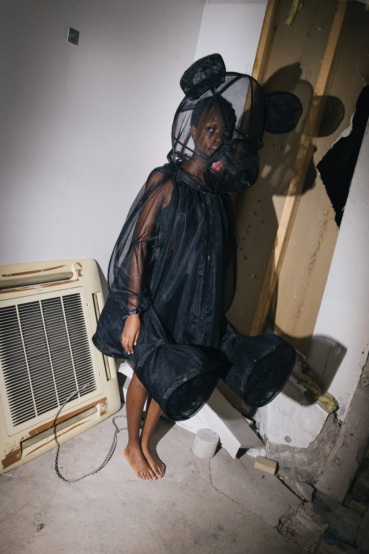 A fashion portrait of a black woman wearing a transparent black teddy bear costume and a shadow of the teddy bear on the wall