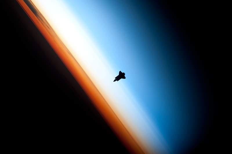 Photo from the International Space Station showing the silhouette of the Space Shuttle "Endeavour" against a colourful backdrop of the layers of the atmosphere: orange, then white, then blue deepening to black.