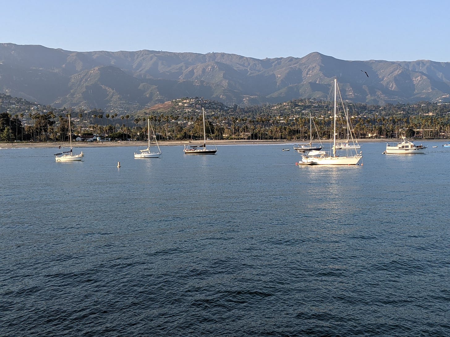 photo of several small sailboats anchored in calm blue water, with a town and rugged mountains in the background