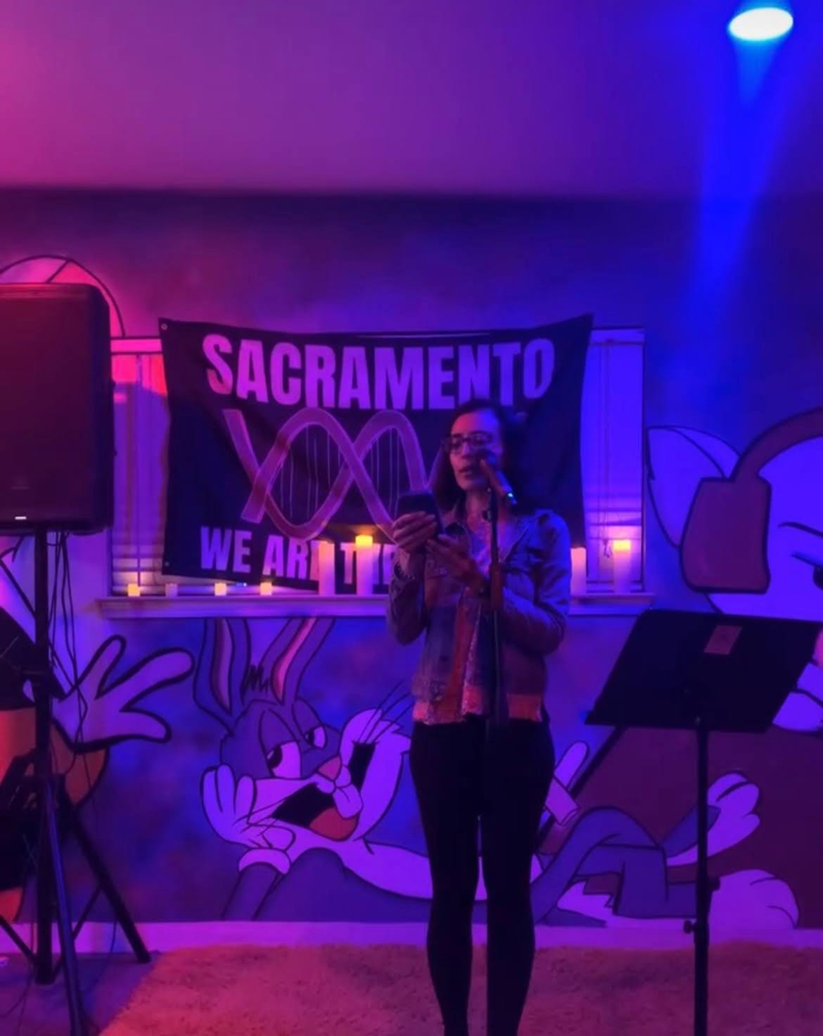 A woman with short hair and glasses stands in front of a microphone and reads from her phone. She's in a room dimly lit with purple and blue lighting.