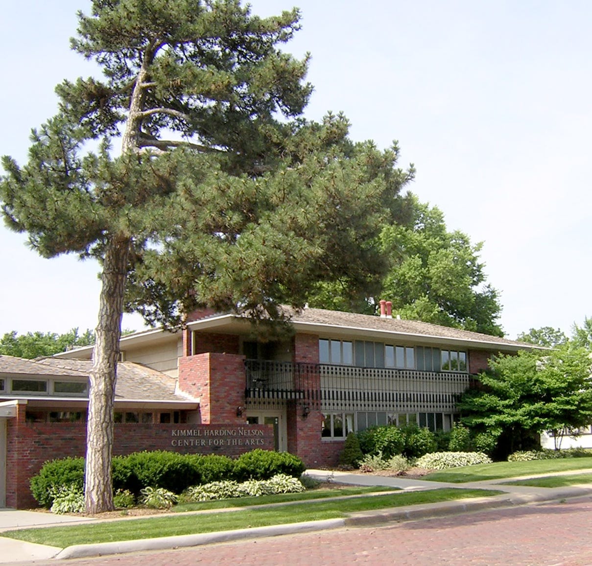 Image of the KHN Center for the Arts, a mid-century brick building with a second story balcony. A large tree is in the foreground of the photo.