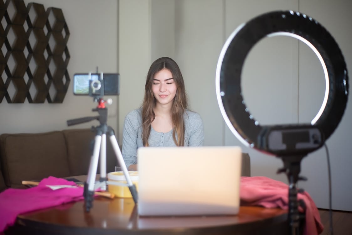 A white woman with brown hair is seating on a brown couch against a white wall. In front of her, on a brown table, are a laptop, a ring light, and a camera on a tripod along with other vlogging tools.