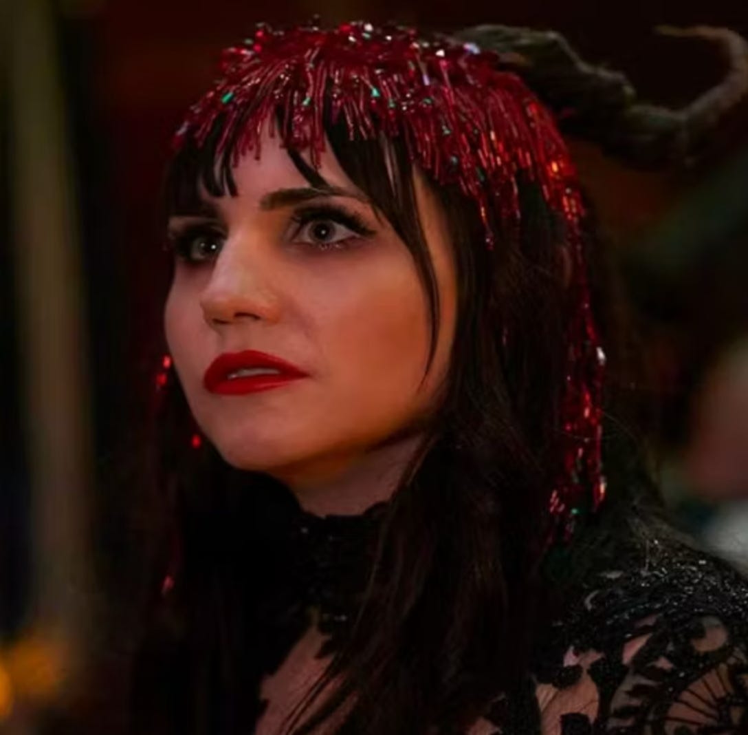 Nadja from What we Do in the Shadows, a white, brown haired vampire who is wearing a glittery red headdress in this picture