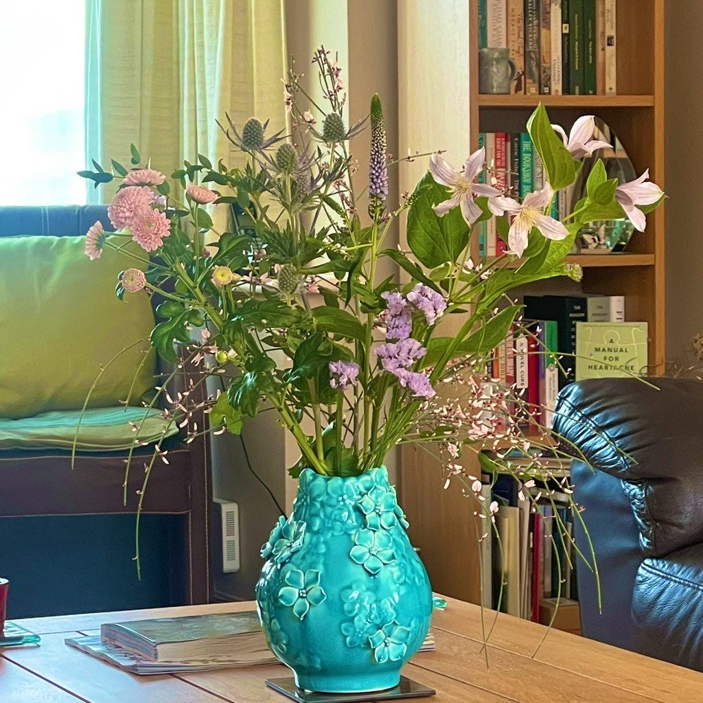 A turquoise vase covered with sculpted flowers sits on a coffee table in a living room. In the vase is a flower arrangement containing 6 stems, each one different. 