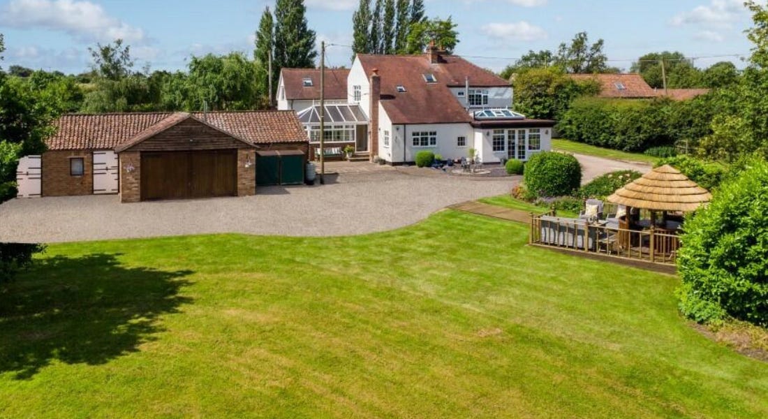 A white house with a red brick roof next to a light brown brick barn, with sprawling green lawns in front and a wooden gazebo area on these