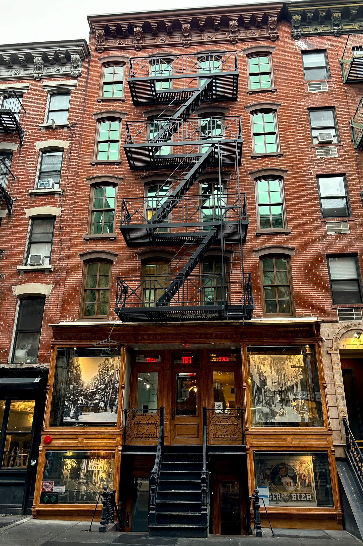 A five-story red brick apartment building with a black fire escape along the facade. The basement level window has a large poster of an ad for lager bier with a cheerful man toasting with a stein. The first floor has a photo of a nearby alley filled with laundry from the turn of the century.