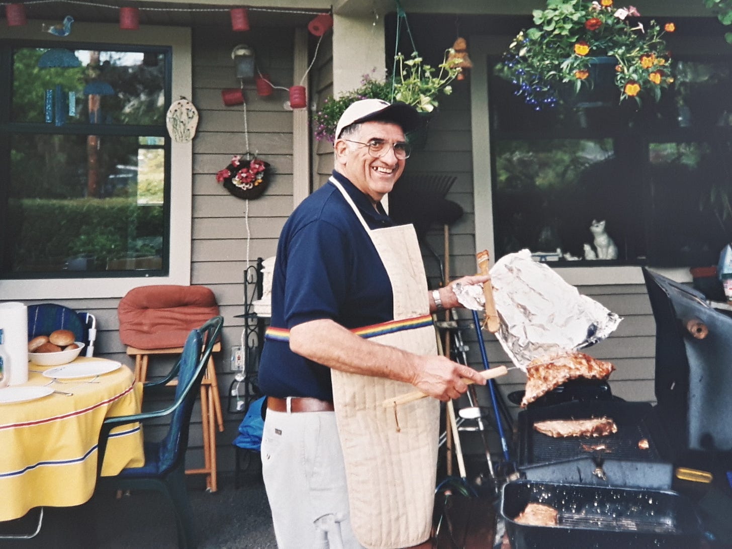 Man in a navy shirt, baseball cap and apron barbequeing ribs on a patio. 