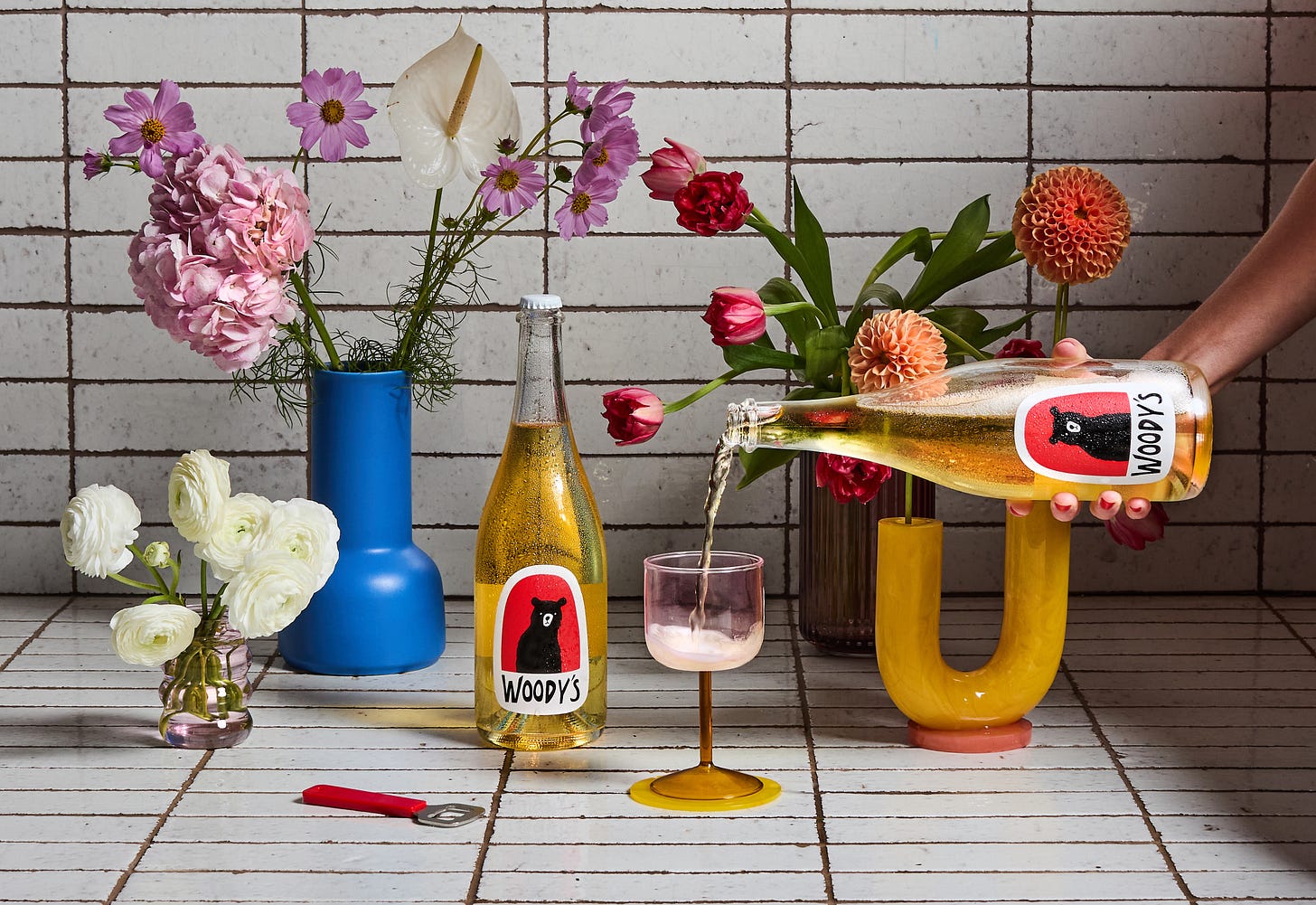 Flowers on a tile counter, bottle of Woody's wine, another bottle of Woody's wine being poured into a pink and amber wine glass.