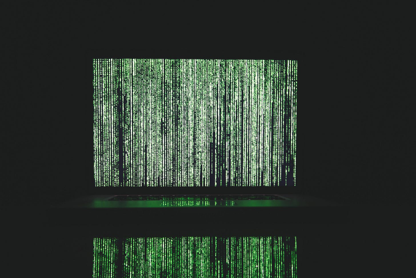 A wall of green and white computer code fills a laptop screen against a pitch-dark background. The code is reflected against the keyboard and, somehow, below the table surface.