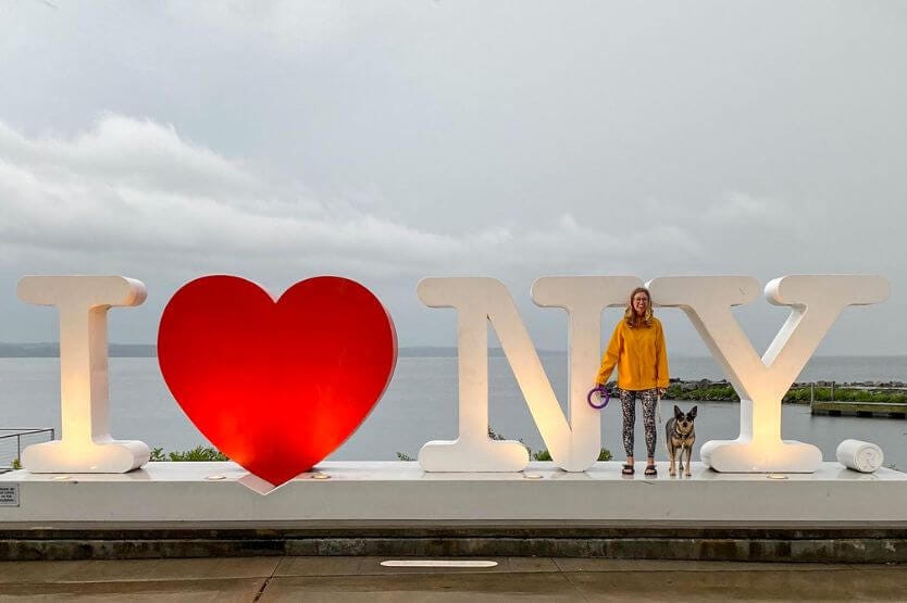 Scout, a blue heeler, and I, a young woman in a yellow raincoat, pose between the N and Y in an I Heart NY sign near the Finger Lakes in upstate New York