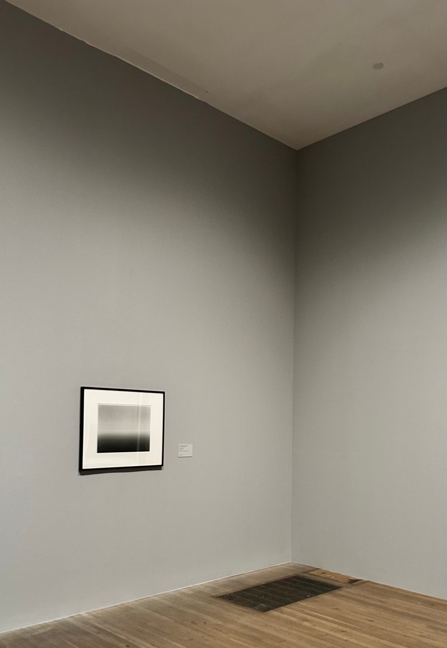 Photo of a dark grey artwork framed with with border hanging in a gallery with grey walls and wooden floor.