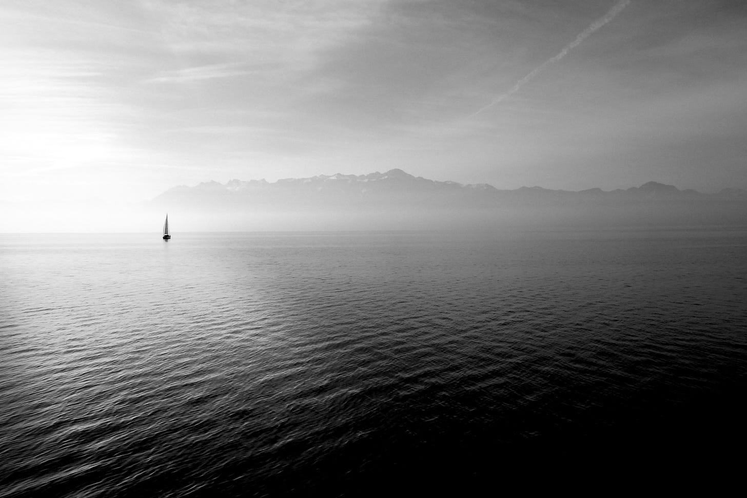distesa di mare con una barca a vela a sinistra e sagome di nuvole sullo sfondo, in bianco e nero