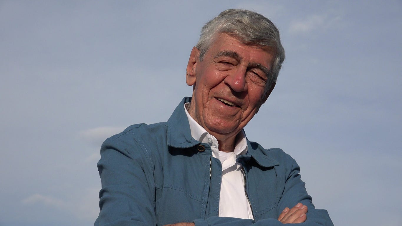 Smiling white man against light blue background with graying hair in blue jacket and white, open-color shirt. His arms are folded, eyes closed.  