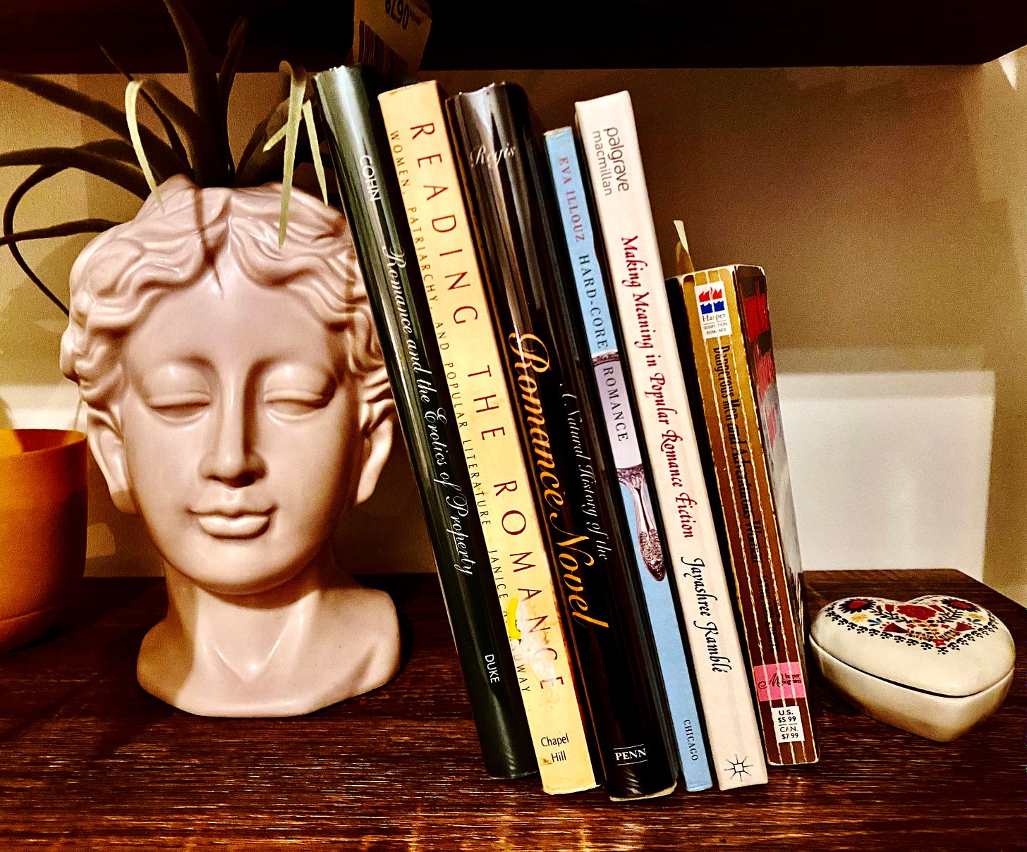 Photo of a stack of romance scholarship books with a bust holding a plant on a bookshelf next to a ceramic heart container.