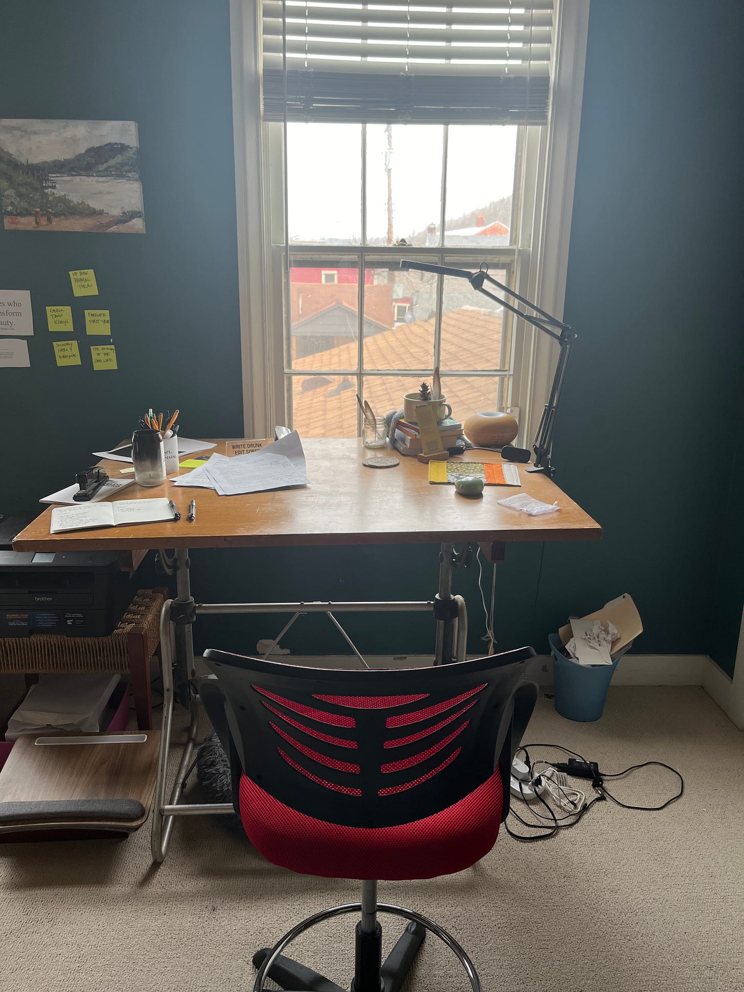 A drafting table sitting in front of a window with the red chair and a view of buildings out the window