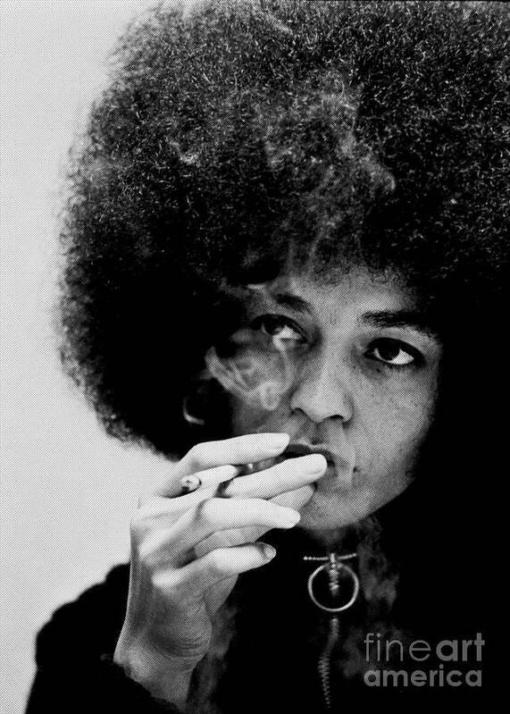 A black and white photo of Angela Davis, activist, writer, and author, smoking a cigarette. She is staring off into the distance. 