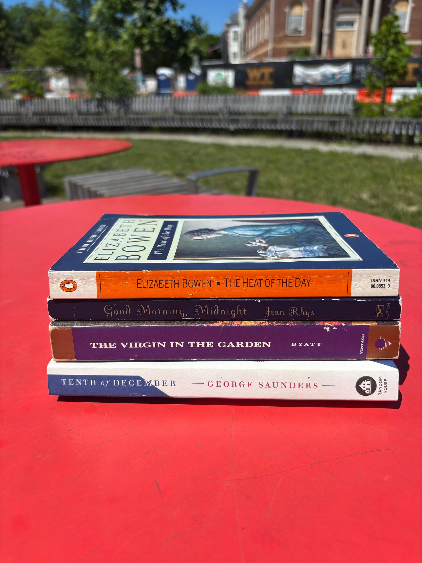 Stack of books (The Heat of the Day by Elizabeth Bowen, Good Morning, Midnight by Jean Rhys, The Virgin in the Garden by A.S. Byatt, and Tenth of December by George Saunders) on top of a red table