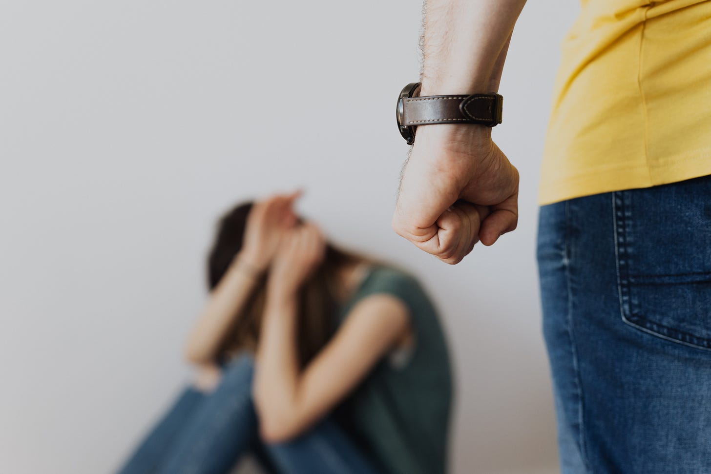 a person wearing a bulky watch, yellow shirt and jeans, with a clenched fist is facing a scared person who is curled agaist the wall with their hands up in a defensive posture