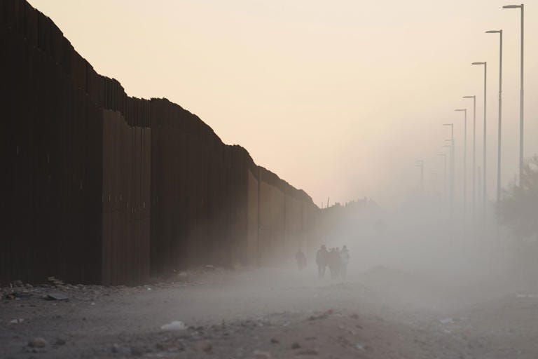 Migrants walk along the US-Mexico border fence in Lukeville, Arizona, US, on Tuesday, Dec. 12, 2023. An influx of migrants crossing the border unlawfully around remote Lukeville, Arizona, has overwhelmed US border officials causing them to close the official port of entry in order to direct resources to processing the unlawful arrivals.