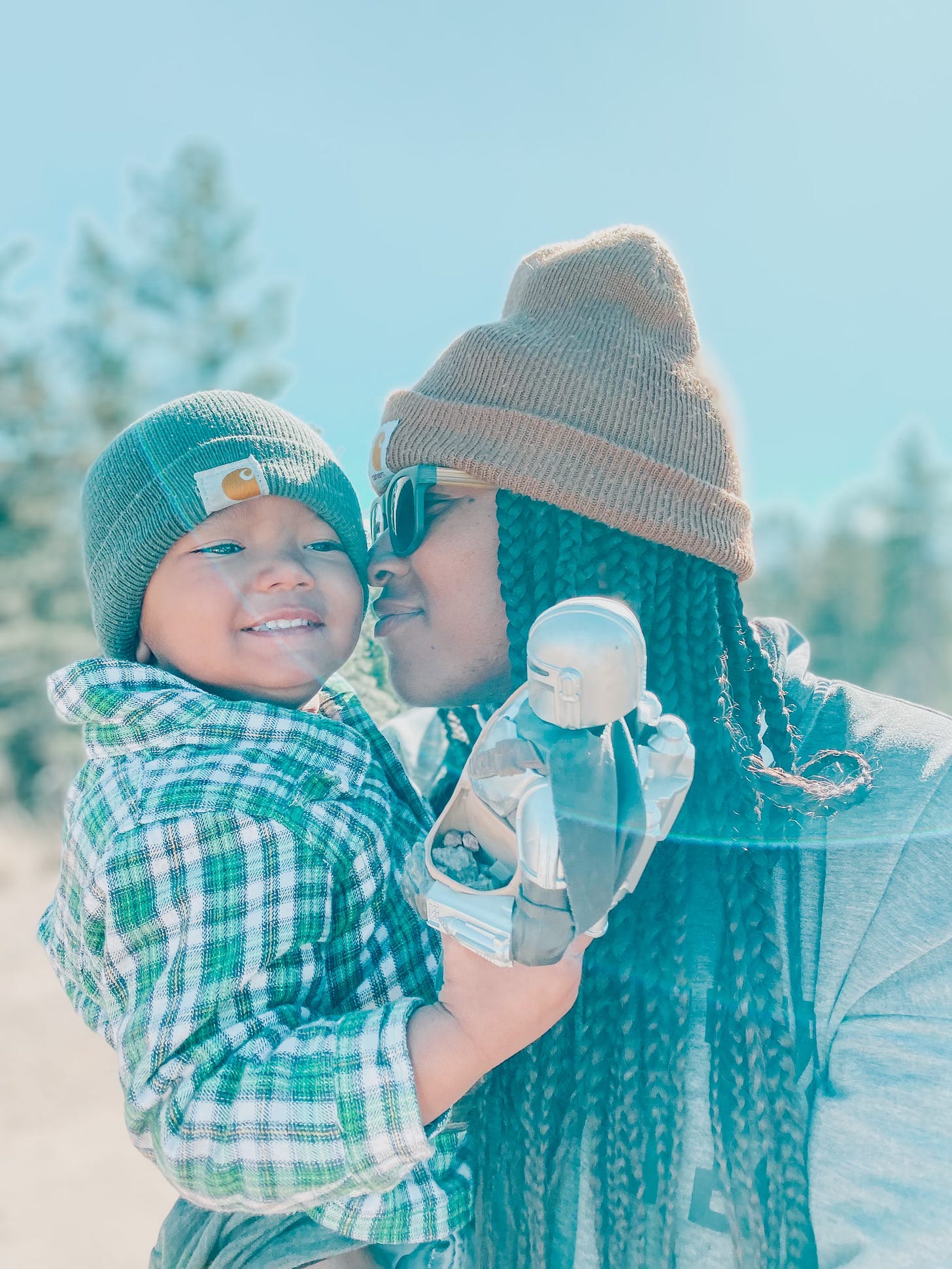A mom and son, standing in the forst. There are tall ponderosa pine trees. Blue skies. The mother holds her 2-year-old son in her arms—nuzzling his cheeks as he smiles.