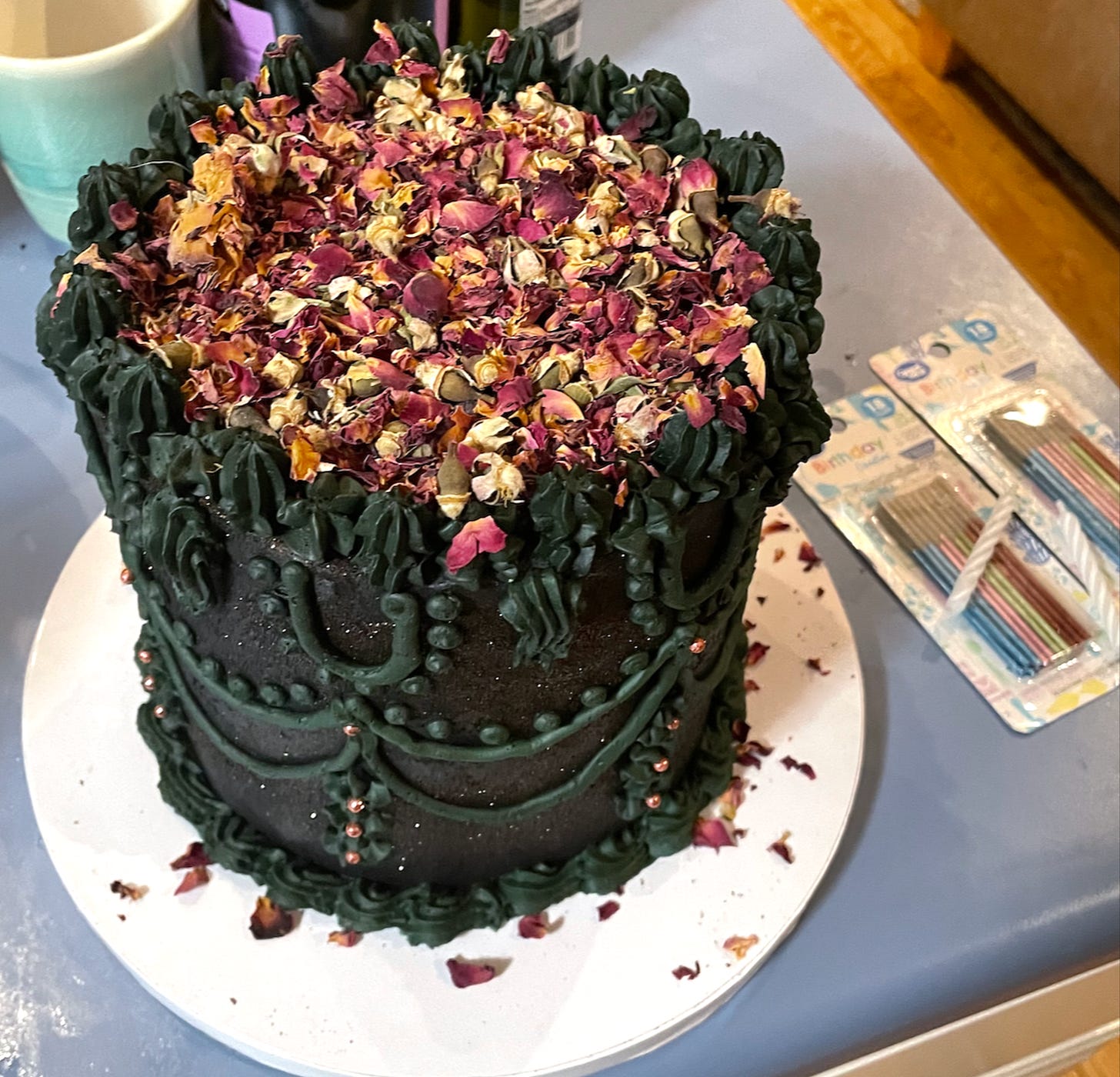 A photo of a handmade tall round birthday cake, iced with black swirly icing on the sides and topped with dried flower petals.