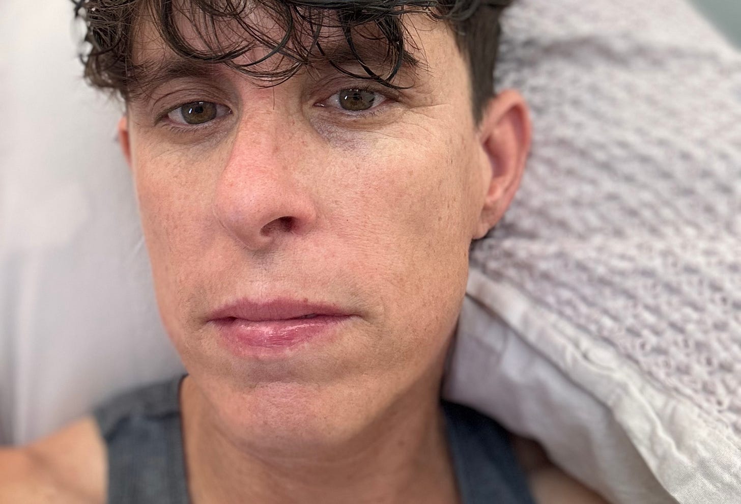A close-up selfie of Andrea Gibson lying on a pillow with tousled dark hair. Their expression is neutral, with a hint of fatigue. Their brown eyes are slightly unfocused, and their skin has a natural, slightly flushed texture. The background consists of a soft, textured pillow and a blanket.