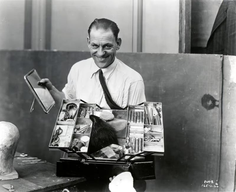 lon chaney, a middle aged white man, grins at the camera. He is holding a train case containing vials, hair, makeup brushes, and other cosmetics tools and materials.