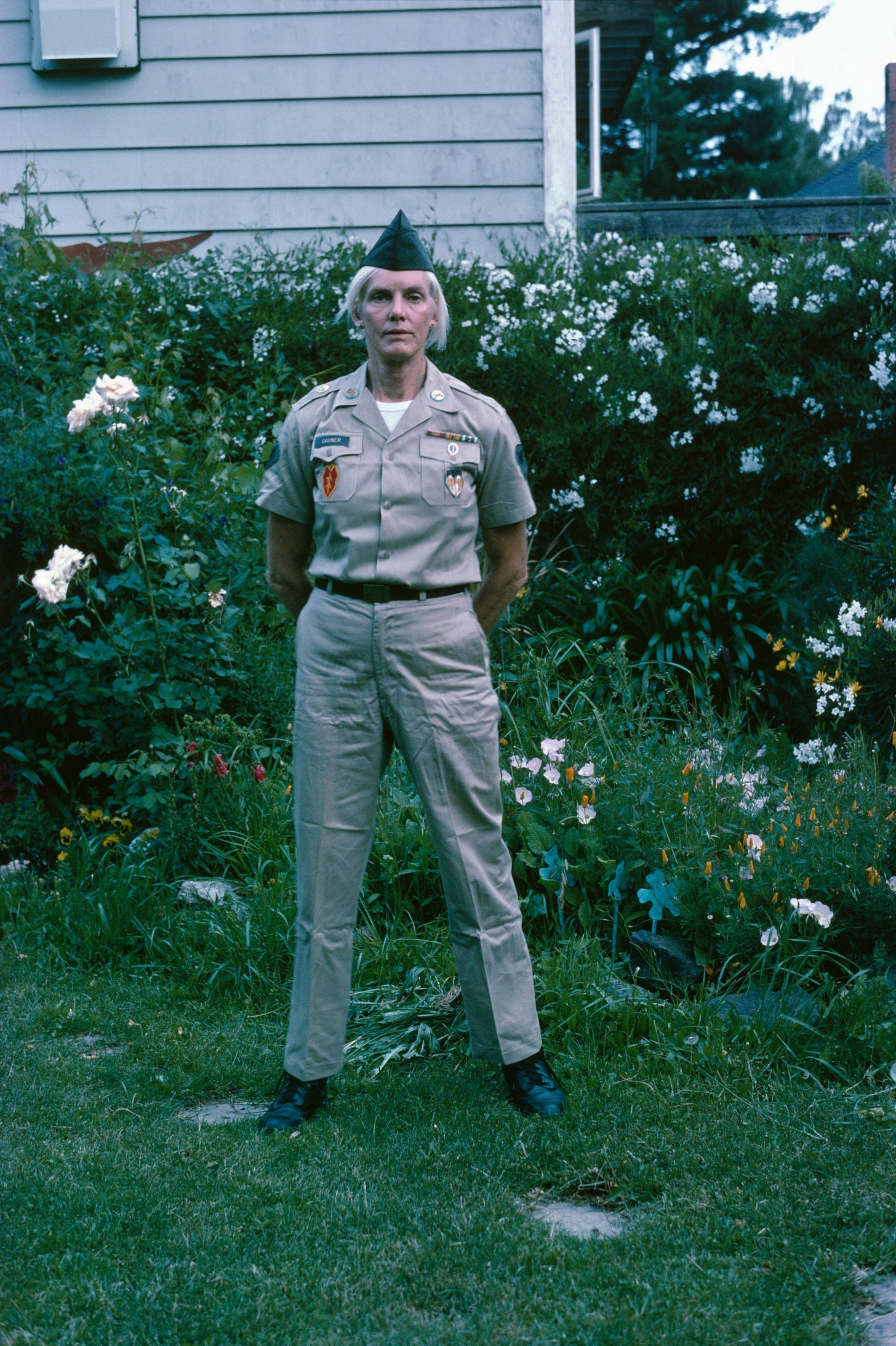 Pippa Garner stands tall and proud in a tan military uniform. She is looking directly into the camera, and is standing in front of a house with a row of shrubs and white flowers in the background. Pippa Garner stands tall and proud in a tan military uniform. She is looking directly into the camera, and is standing in front of a house with a row of shrubs and white flowers in the background.