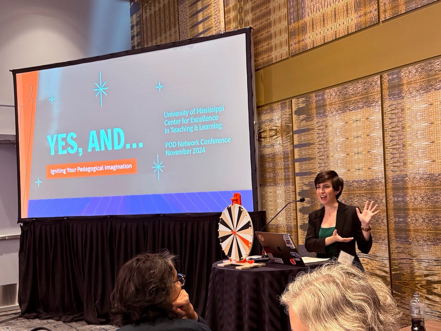 Emily gesturing to the out-of-frame panelists; to the left is a spinning prize wheel and a screen that reads "YES, AND... Igniting Your Pedagogical Imagination."