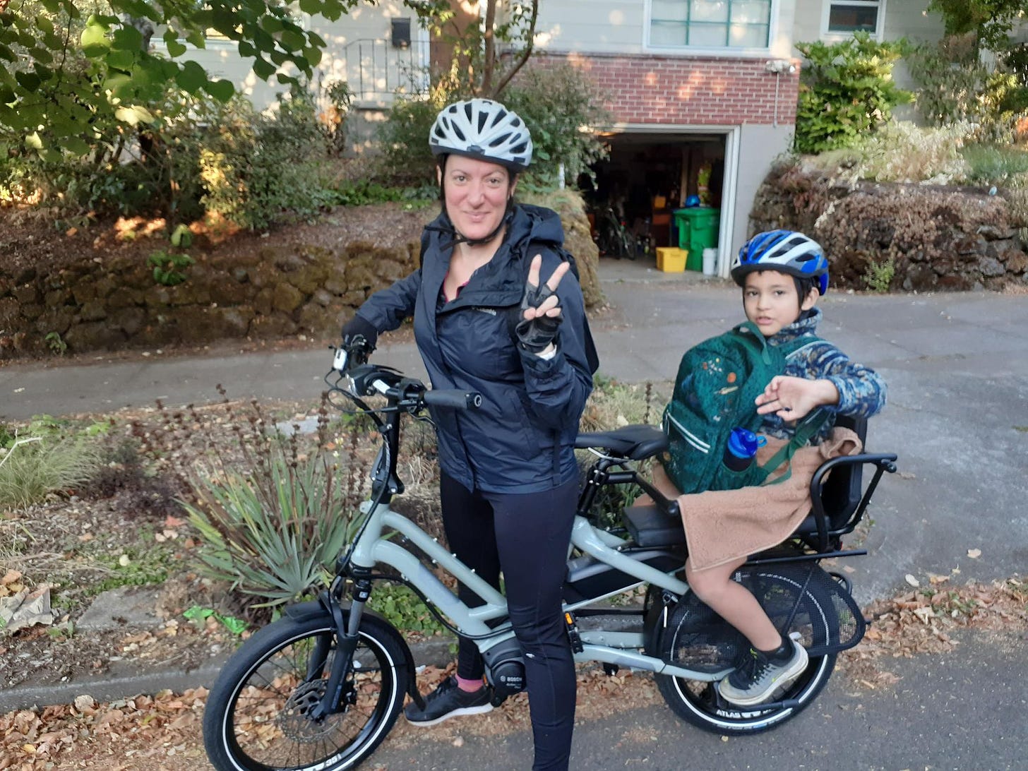 Nicolai is stood over her own cargobike about to depart her house. Her son is on the back seat holding his school bag. 