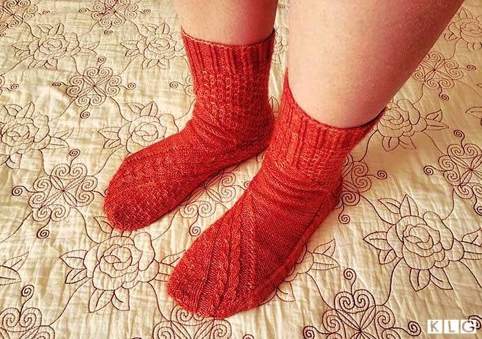 Top view of the two Smaug socks showing the gorgeous cable stitches and dragon scale stitch pattern with ribbed cuff in coral colored yarn